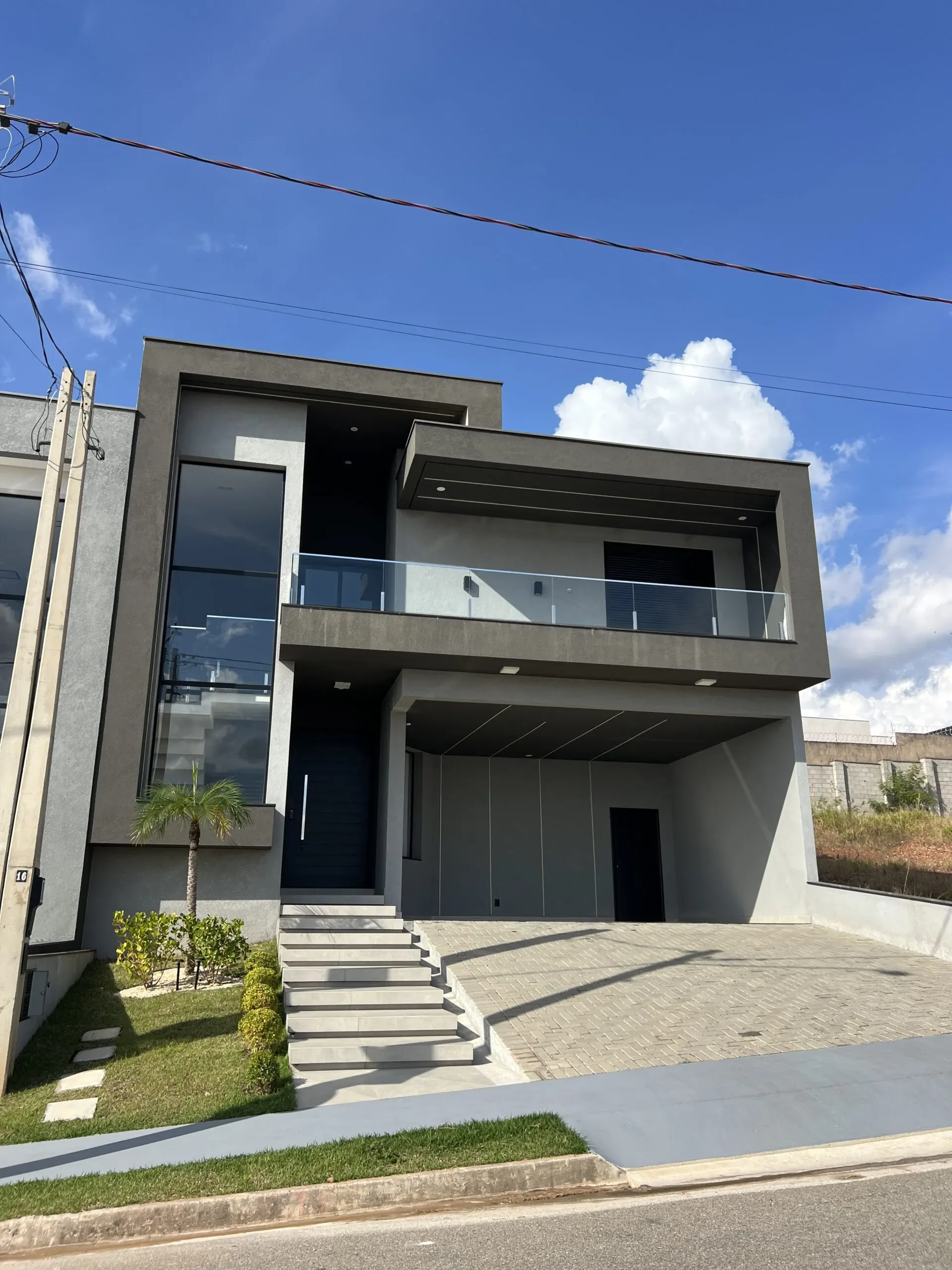 Casa moderna com arquitetura reta, pintura cinza escuro, guarda-corpo de vidro na varanda superior, janela alta à esquerda com esquadria preta e porta de entrada escura.