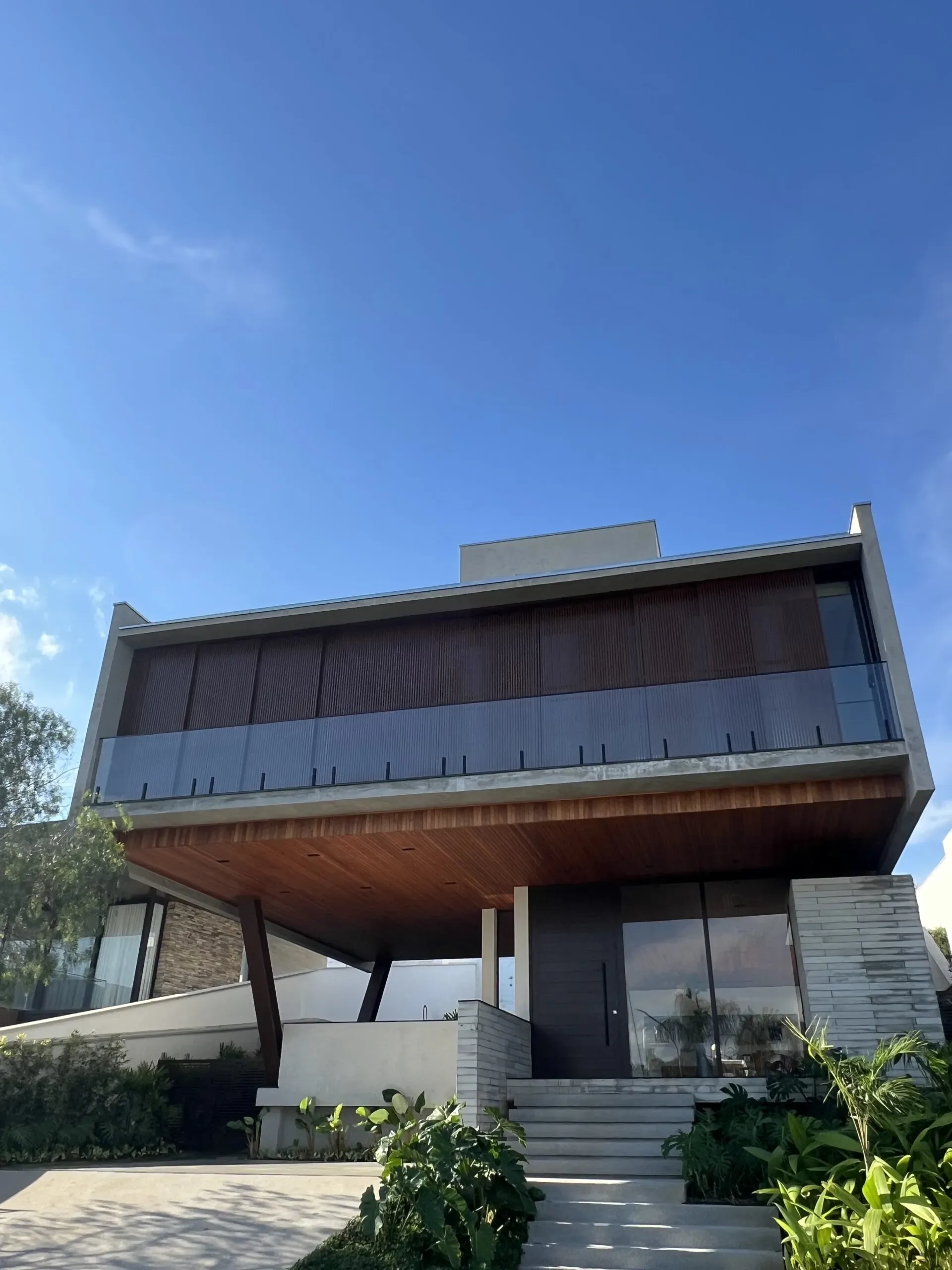 Entrada de residência imponente com porta de giro escura, pé direito alto e varanda superior com painel ripado.