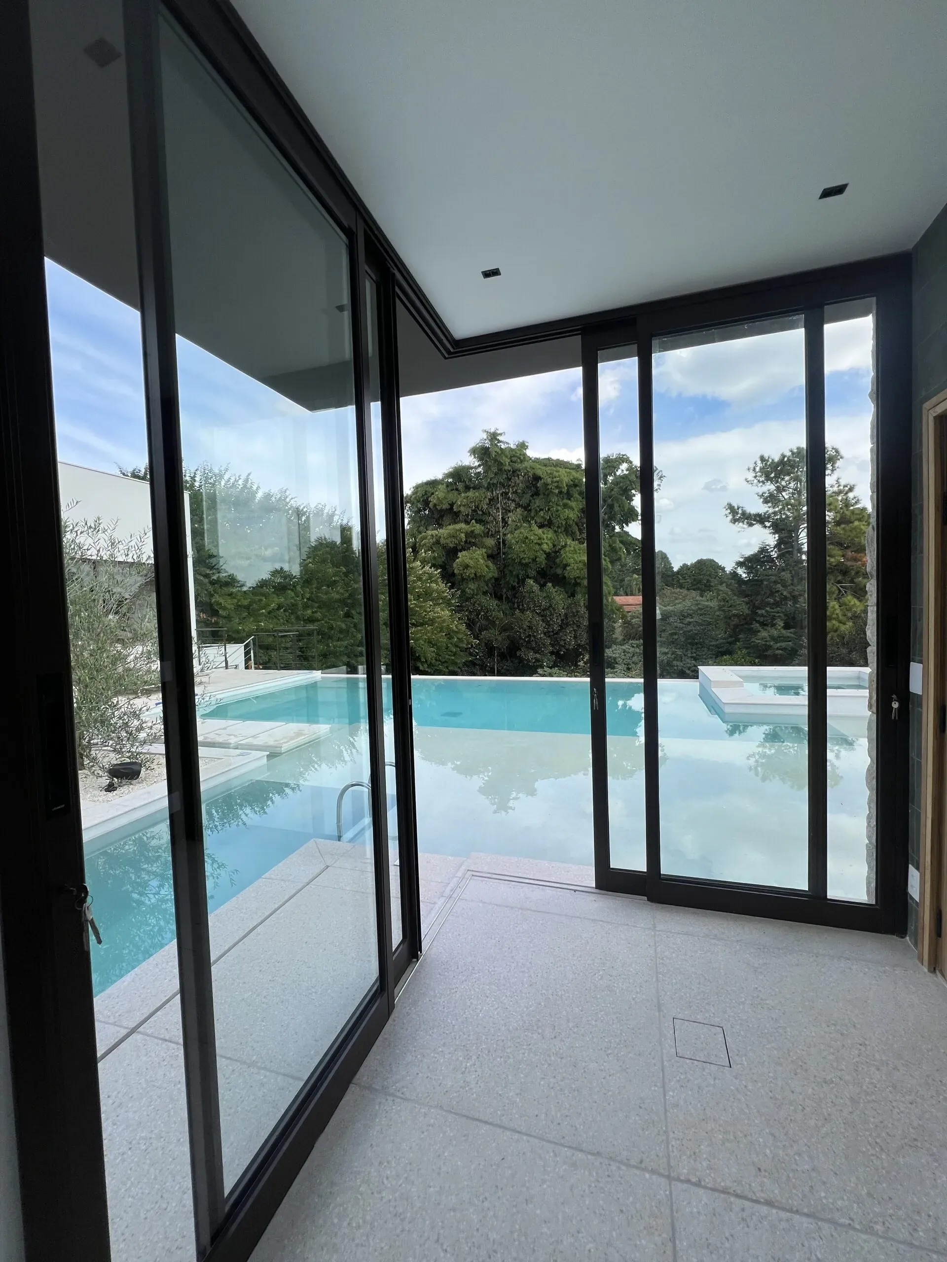 Sistema de esquadrias de correr em alumínio preto em canto de 90 graus abrindo para área de piscina com vista para jardim e arborização.