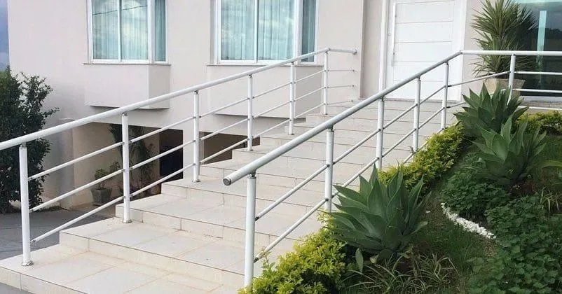 Guarda-corpo tubular em alumínio pintado na cor branca instalado em escada externa de casa, ladeado por jardim com plantas ornamentais.