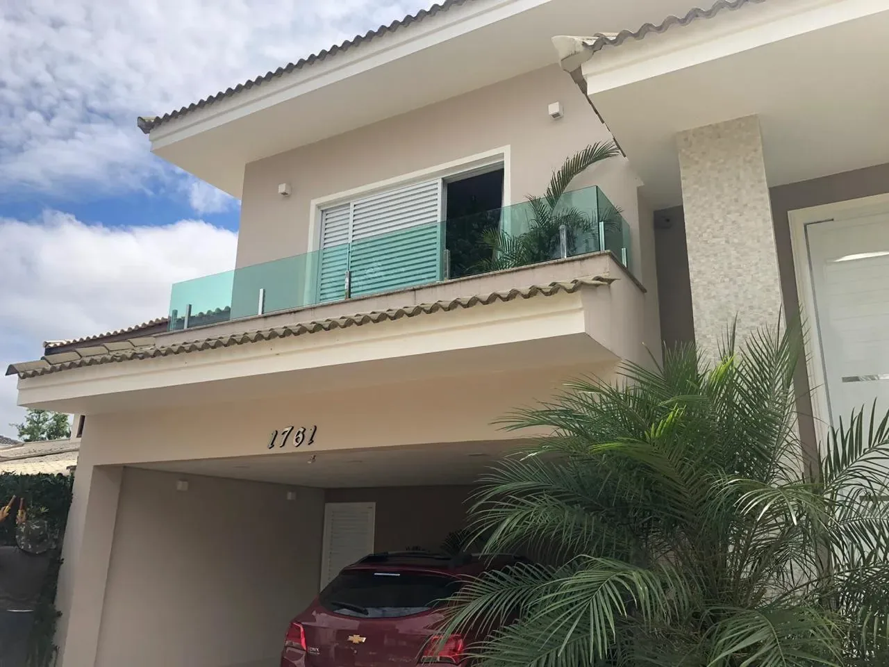 Sacada de residência em condomínio com guarda-corpo de vidro temperado na tonalidade verde, fixado por ponteiras, com palmeira ao fundo e carro na garagem.