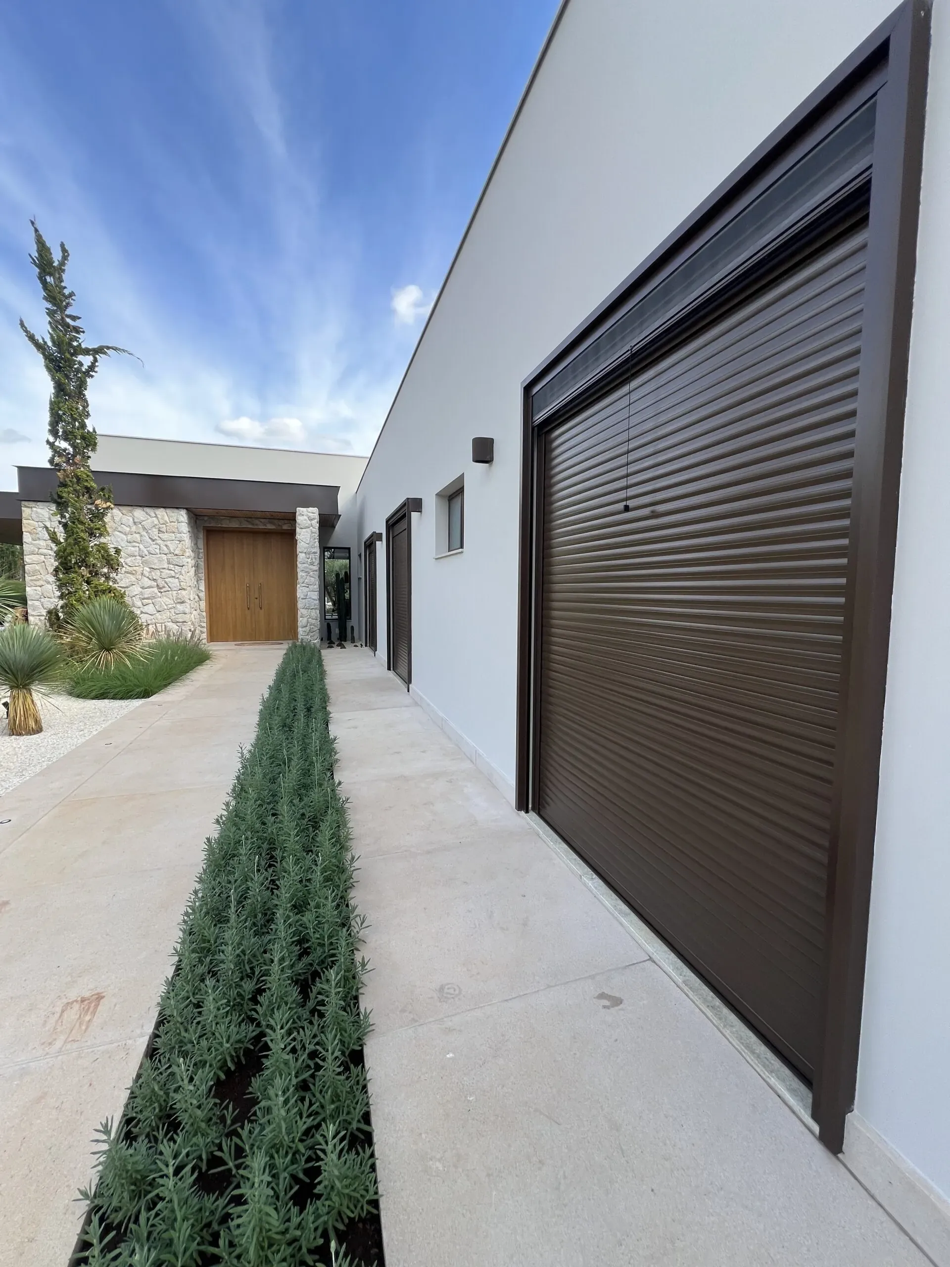 Lateral de residência contemporânea com persiana de enrolar em alumínio marrom escuro e esquadrias combinando, ao fundo porta de madeira maciça.