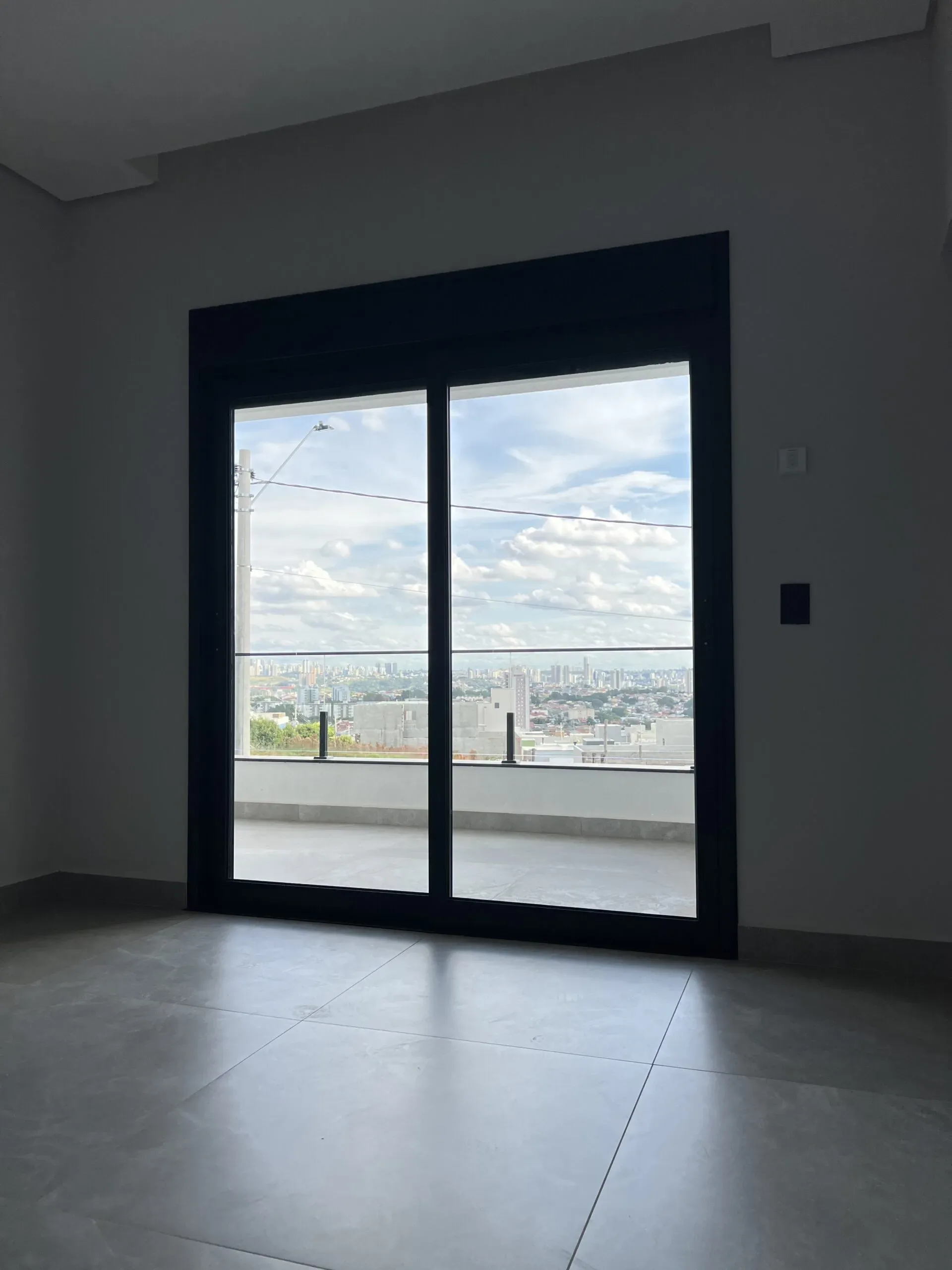 Porta balcão preta de alumínio com persiana levantada e guarda-corpo de vidro ao fundo.
