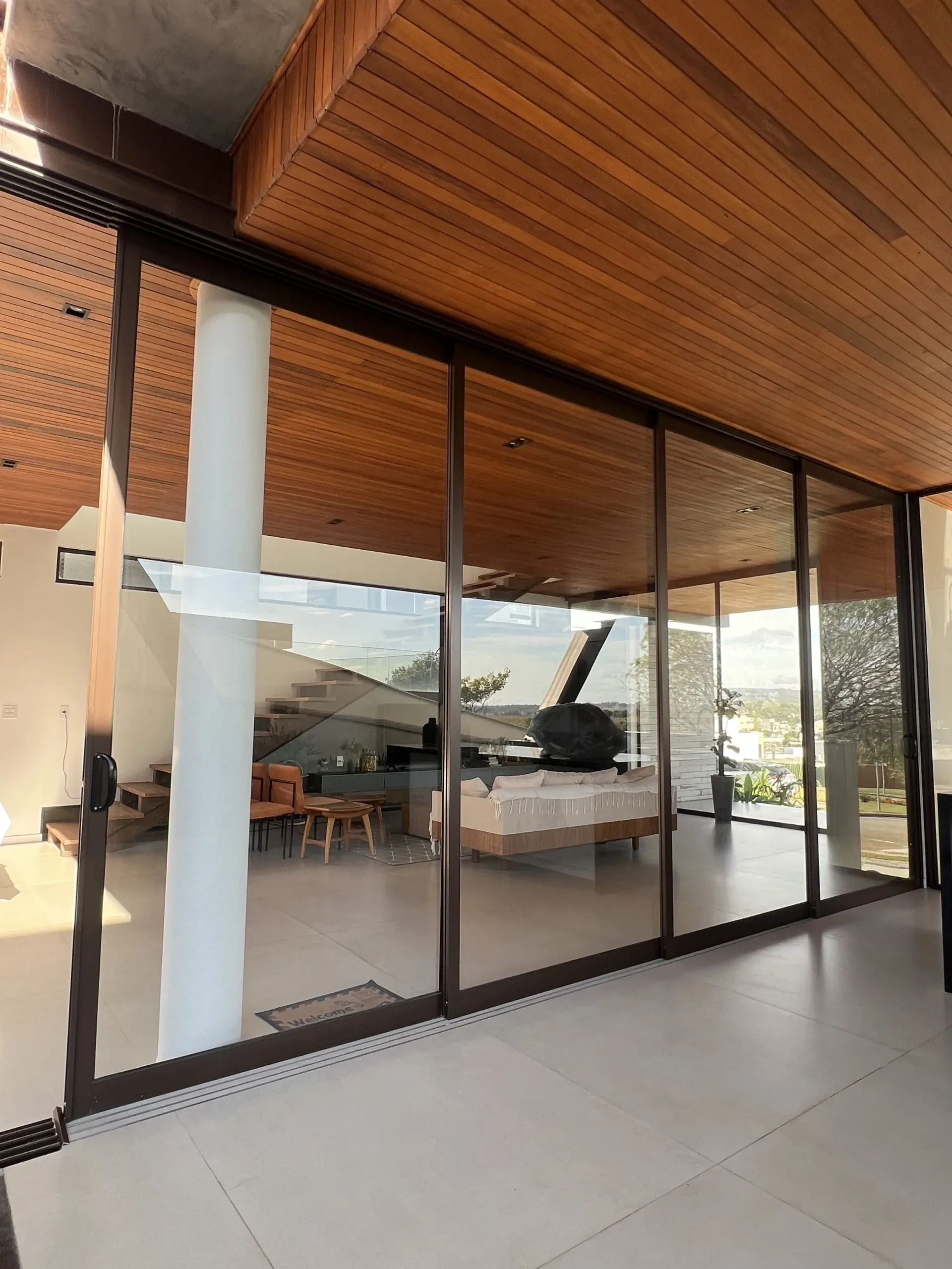 Sistema de porta de correr em alumínio bronze com perfis finos e vidro amplo, revelando sala de estar moderna com escada suspensa e paisagismo externo ao fundo.