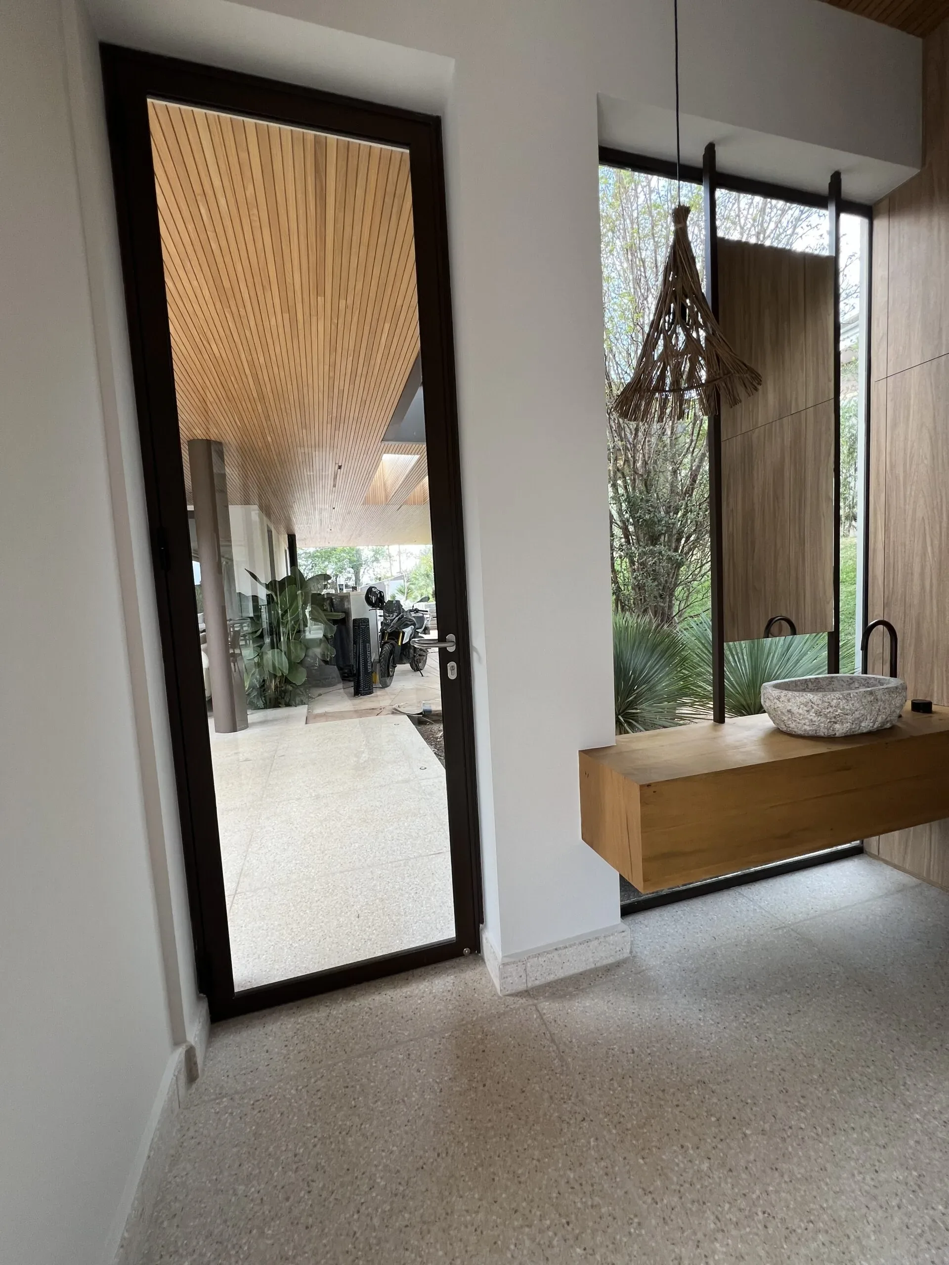 Porta de abrir em alumínio marrom com vidro temperado instalada em ambiente interno moderno, revelando área de garagem coberta com forro de madeira.