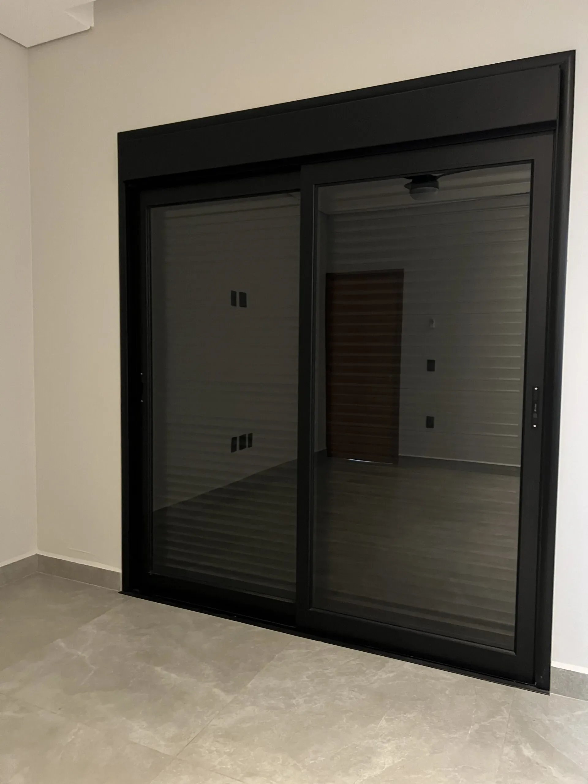 Porta de vidro de correr e persiana de alumínio preta fechada em sala vazia.