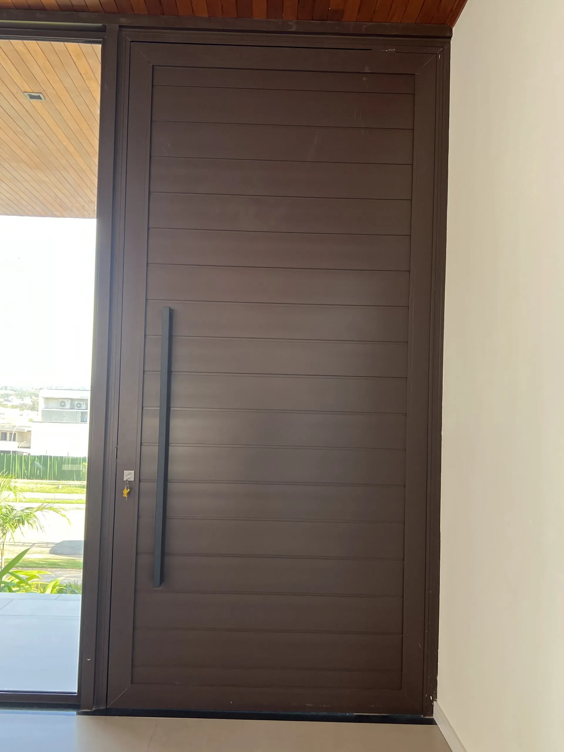 Vista interna da porta pivotante em alumínio marrom com ripas horizontais e puxador slim, enquadrada em vão amplo com parede branca e abertura lateral com vista para área externa.