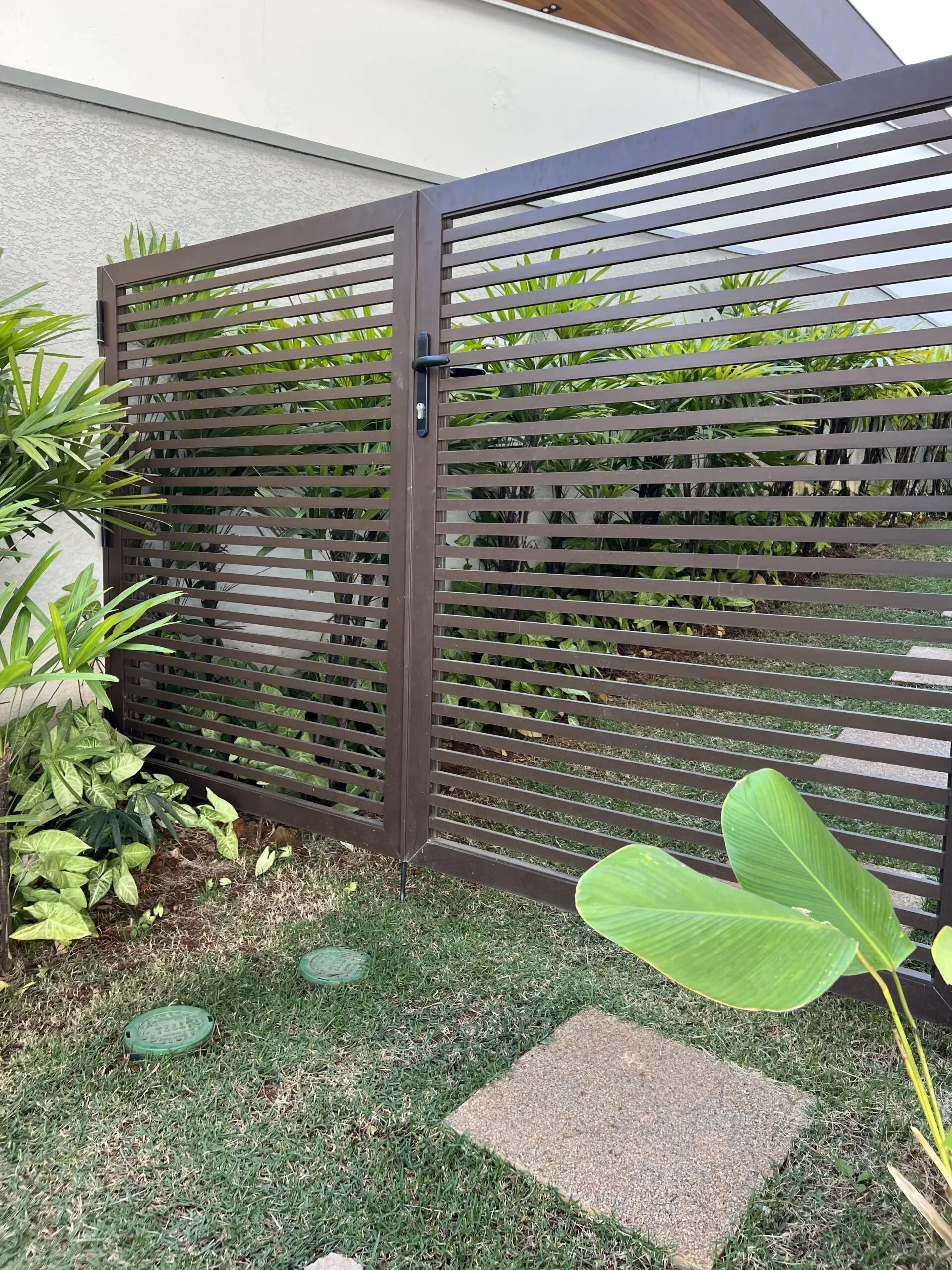 Portão de alumínio em tom marrom escuro com ripas horizontais espaçadas, instalado no jardim lateral de residência com vegetação tropical ao redor.