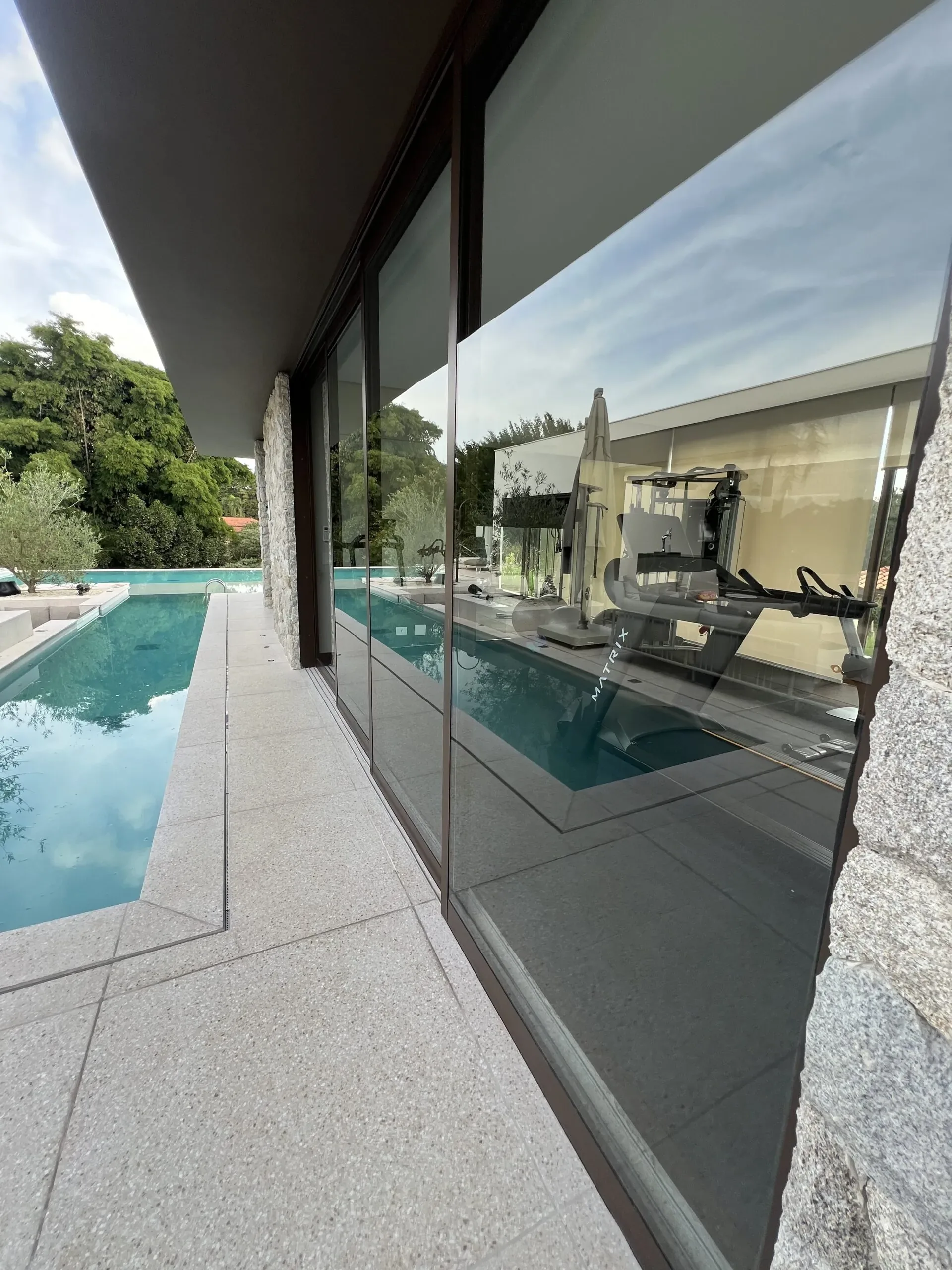 Reflexo no vidro de área com academia, vista para piscina e parede de pedra natural.