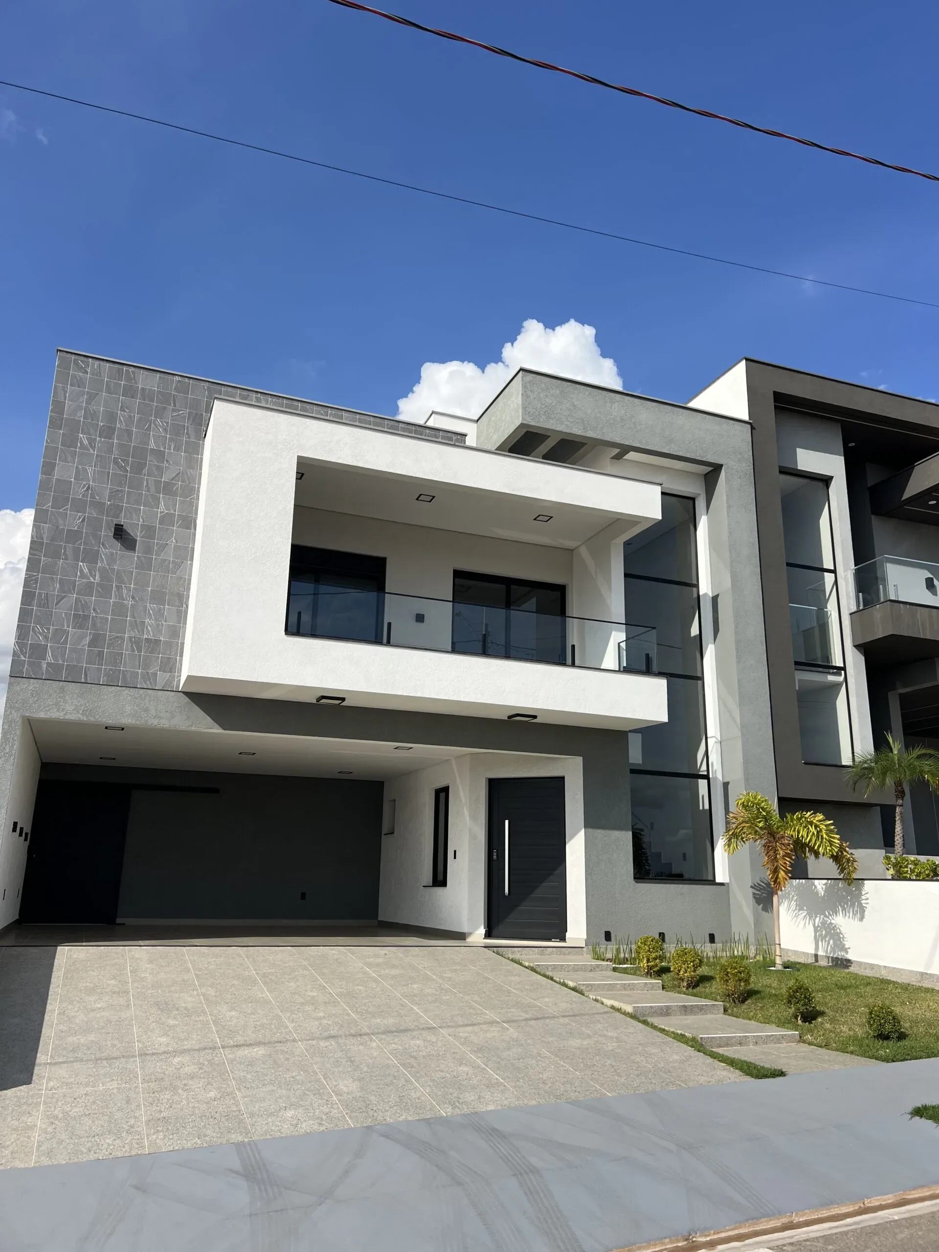 Vista da fachada de residência em estilo moderno, com elementos em cinza e branco, sacada superior protegida com vidro, janela alta à direita e rampa cimentada.