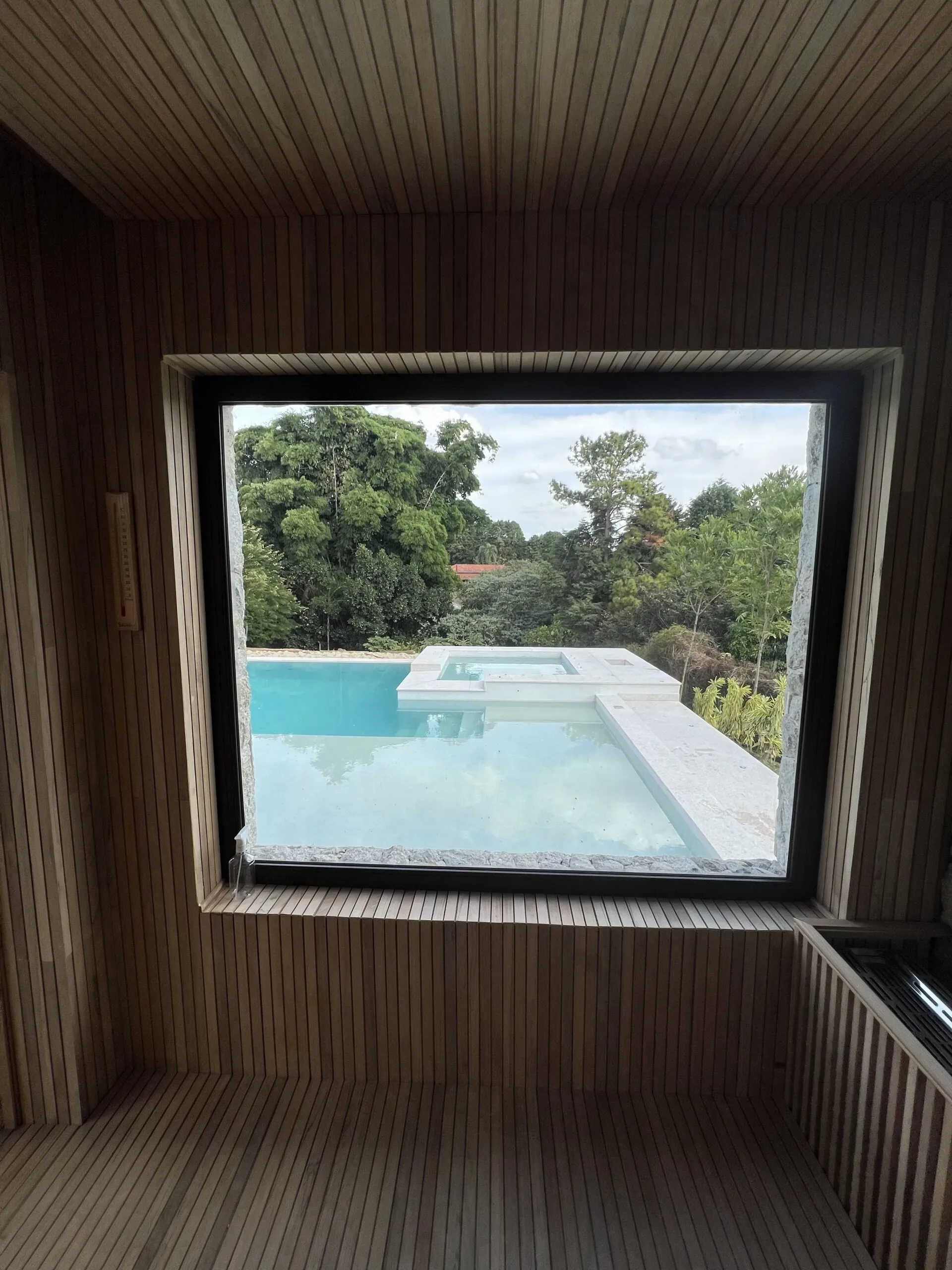 Vista de dentro de sauna com revestimento de madeira através de janela de vidro fixa para piscina e área verde externa
