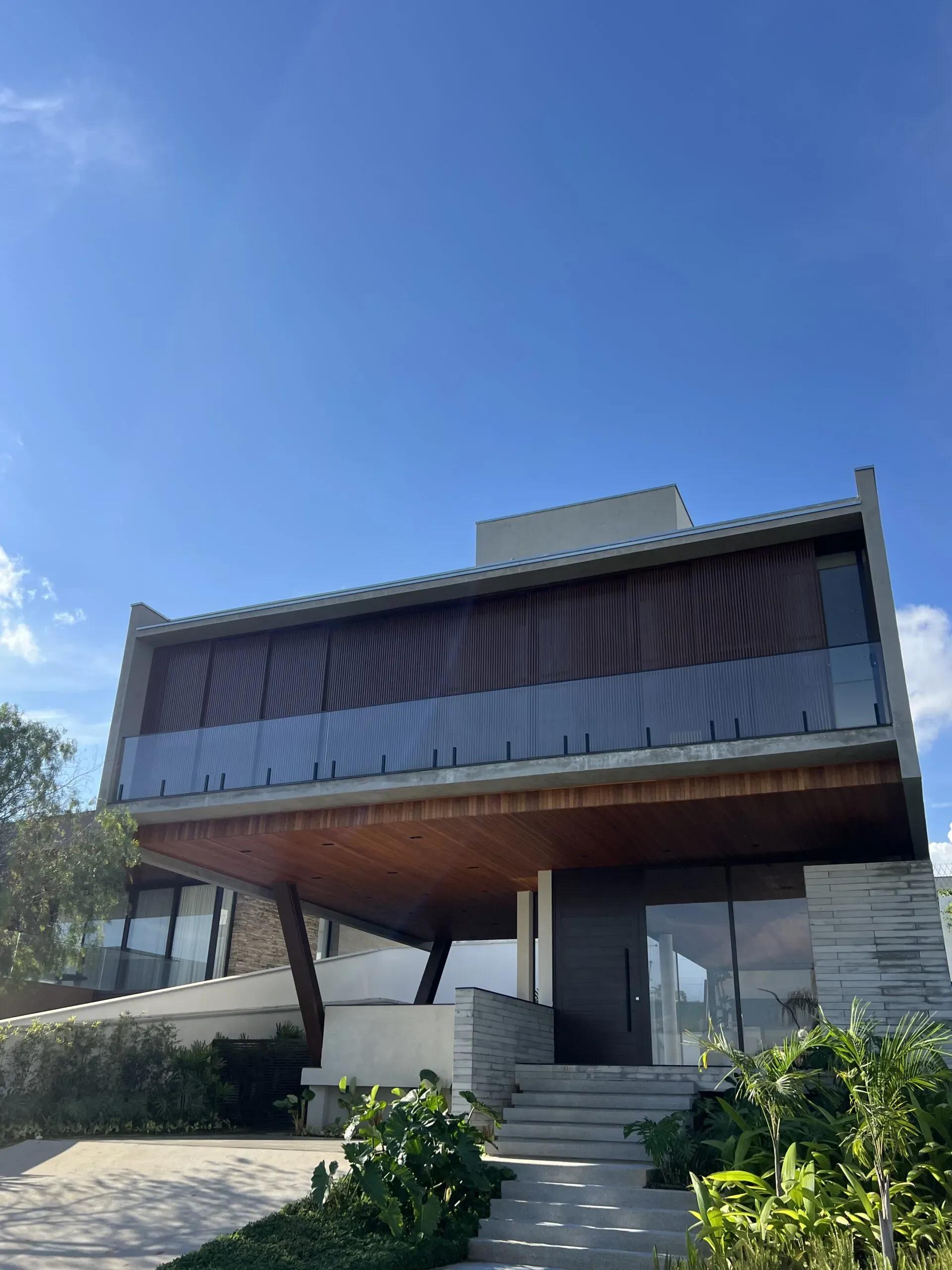 Vista externa da casa com destaque para a escada de acesso, porta de giro escura e brises verticais.