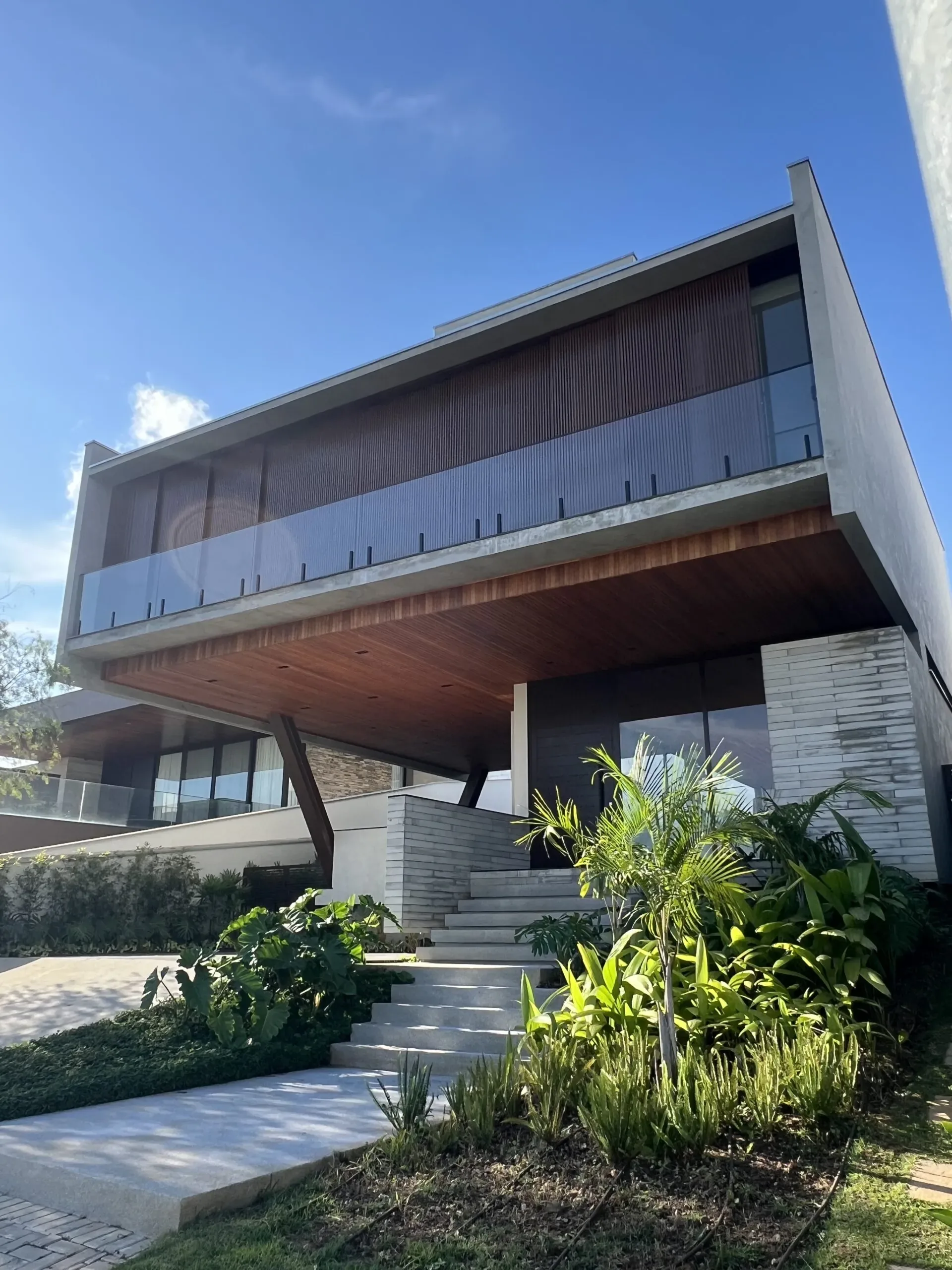 Vista lateral da fachada residencial moderna com esquadrias de alumínio preto e guarda-corpo de vidro, cercada por vegetação tropical exuberante e escadas externas em concreto.